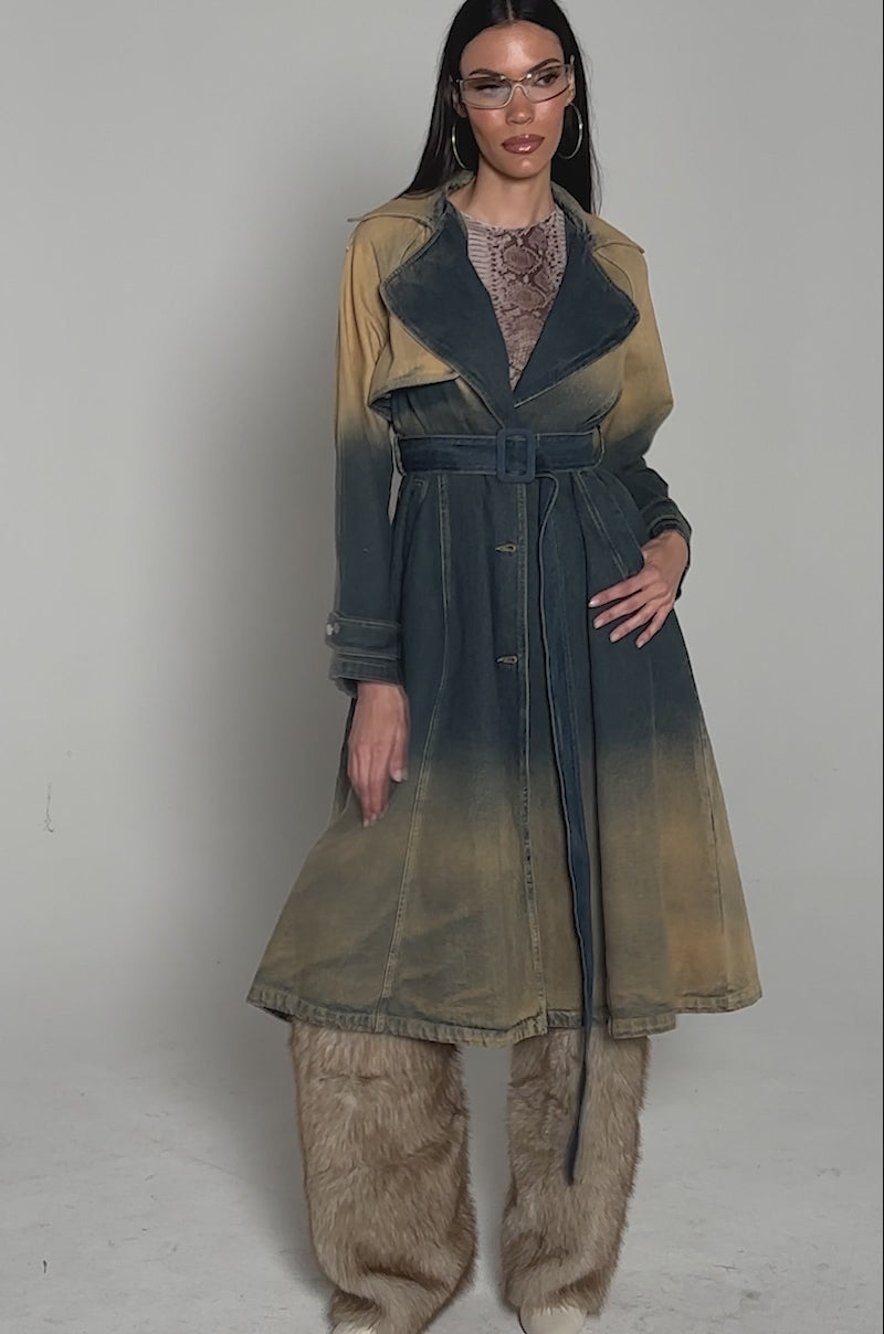 Video of woman modeling the ACID WASHED DENIM TRENCH with tan accents, clear visor sunglasses, large hoop earrings, a patterned top, and fur-trimmed boots stands against a plain light background.