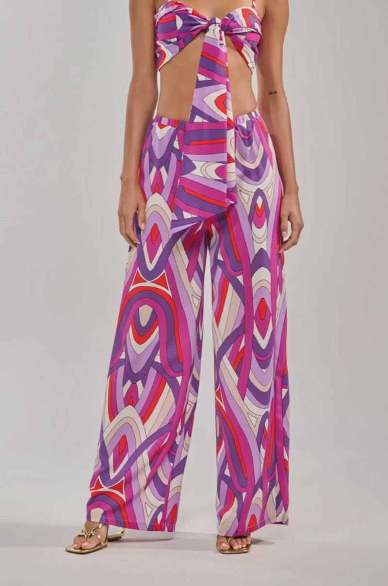 A video of a woman wearing the GIRLY POP BRALETTE, featuring vibrant purple geometric patterns and a front tie, paired with matching high-waisted pants.