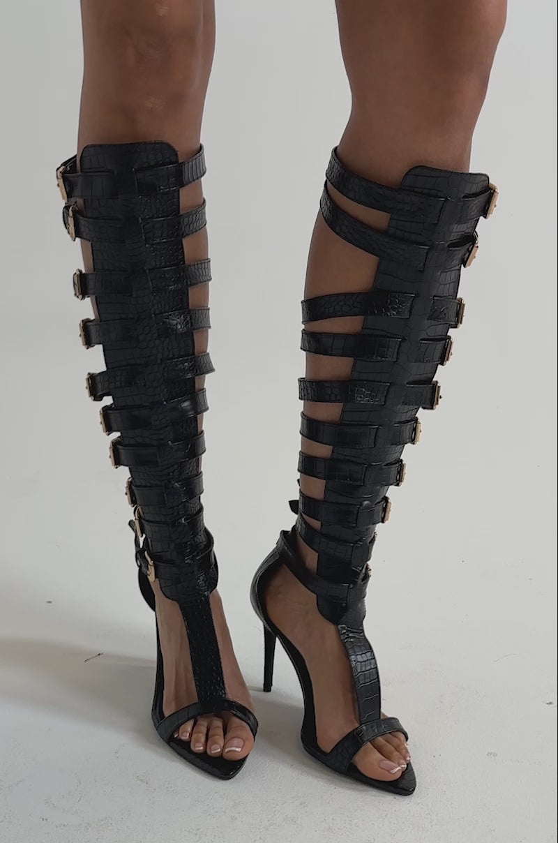 A video of a person wearing AZALEA WANG CHERLLI BLACK CROC OVER THE KNEE SANDALS with multiple gold-buckled straps, and faux leather stiletto heels.