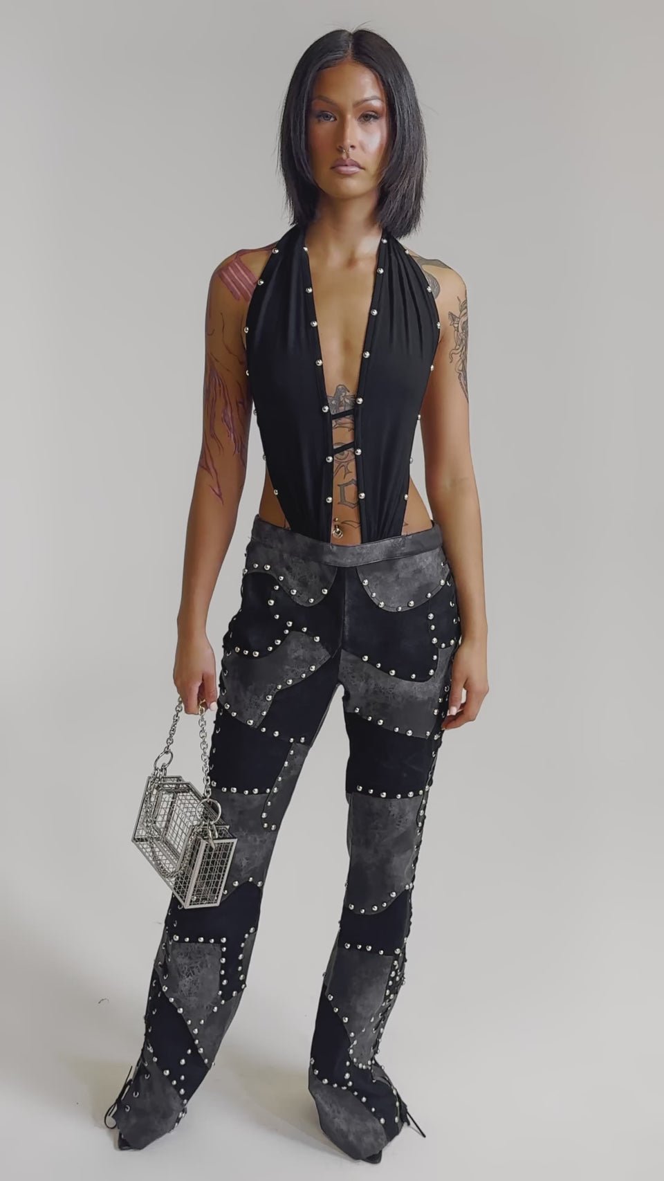 A video of a woman in the ROXY STUDDED SLEEVELESS JUMPSUIT, featuring black studded details and lace-up accents, holding a small silver handbag against a plain light gray background.