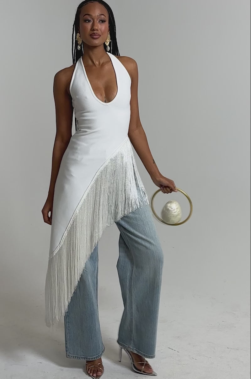 A video of a woman wearing the TAKE ME OUT FRINGE TOP—white with silver diagonal fringe—paired with wide-leg light blue jeans, gold sandals, and a small gold handbag.