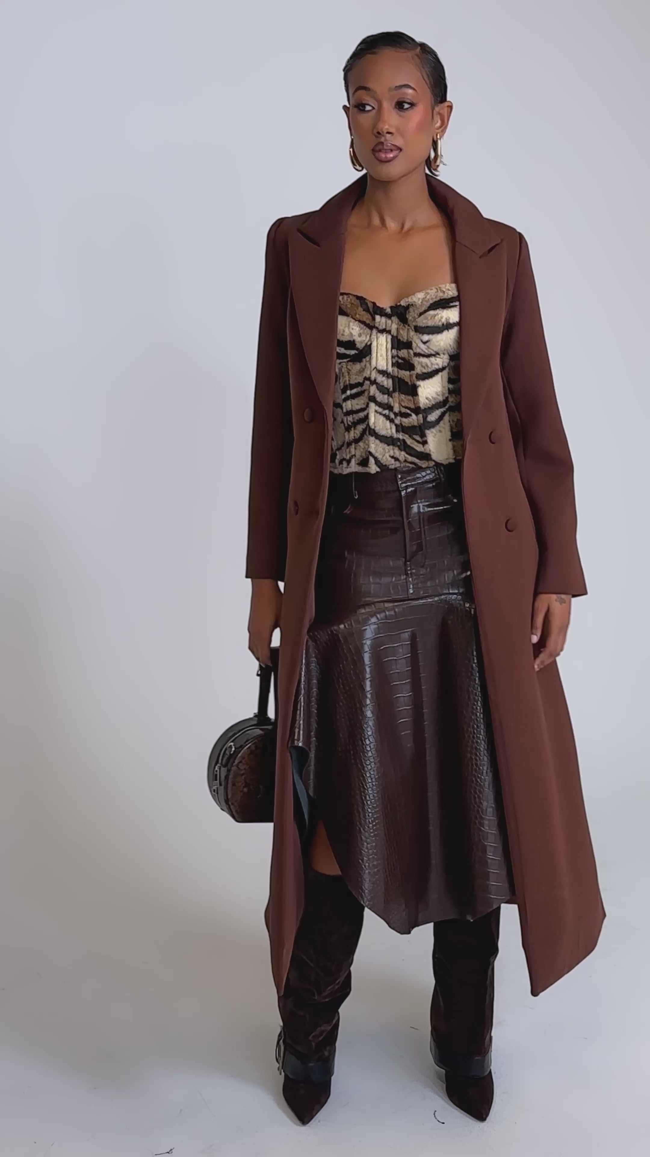 A video of a woman in the COCOA BUTTER SCUBA TRENCH IN BROWN, layered over a tiger-print top, an asymmetrical dark brown leather skirt, and knee-high boots. 