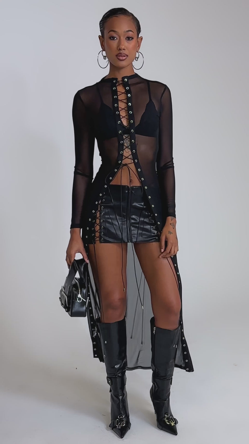A video of a woman in the SEE THROUGH ME BLOUSE, styled with a black bra, black lace-up mini skirt, knee-high black boots, large hoop earrings, and a small black handbag.
