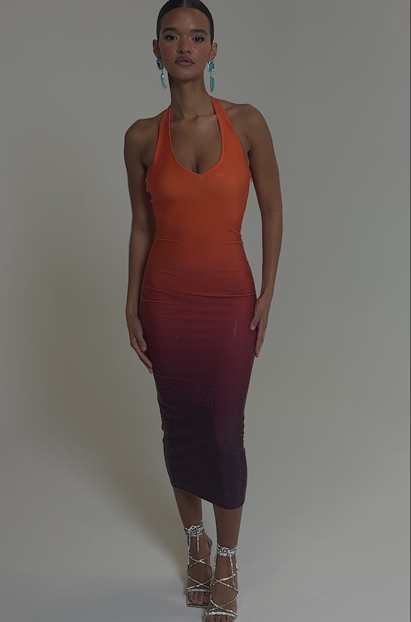 Video of woman modeling the DUSK TILL DAWN RHINESTONE MIDI DRESS IN ORANGE with a sleeveless, halter-neck ombre design fading to pink and purple, she pairs it with blue statement earrings and strappy silver heels.