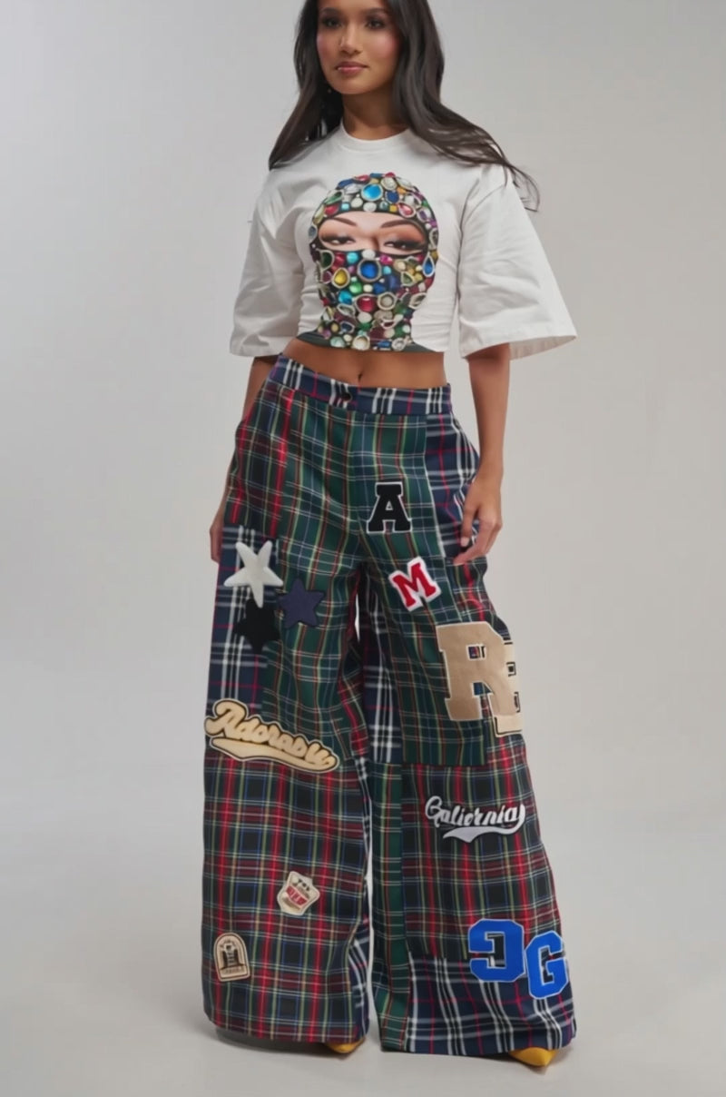 Video of woman modeling the FACE CARD GRAPHIC CROPPED TEE, which features a jeweled ski mask graphic. She pairs it with plaid pants, hands in pockets, and poses against a plain background for an effortless streetwear look.