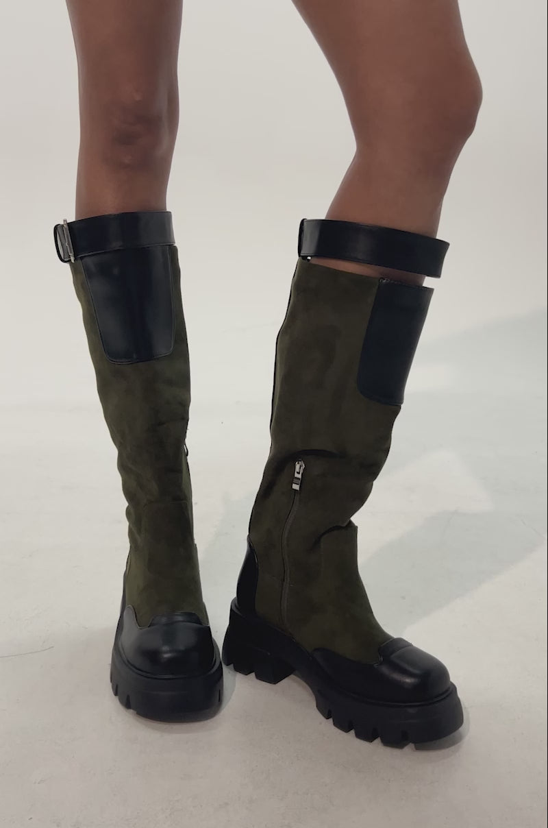 Video of woman modeling the AZALEA WANG DALILA OLIVE FAUX SUEDE FLAT BOOT—olive green knee-high faux suede boots with chunky black soles, black toe and heel accents, a side zipper, and top buckle—paired with a plaid skirt.