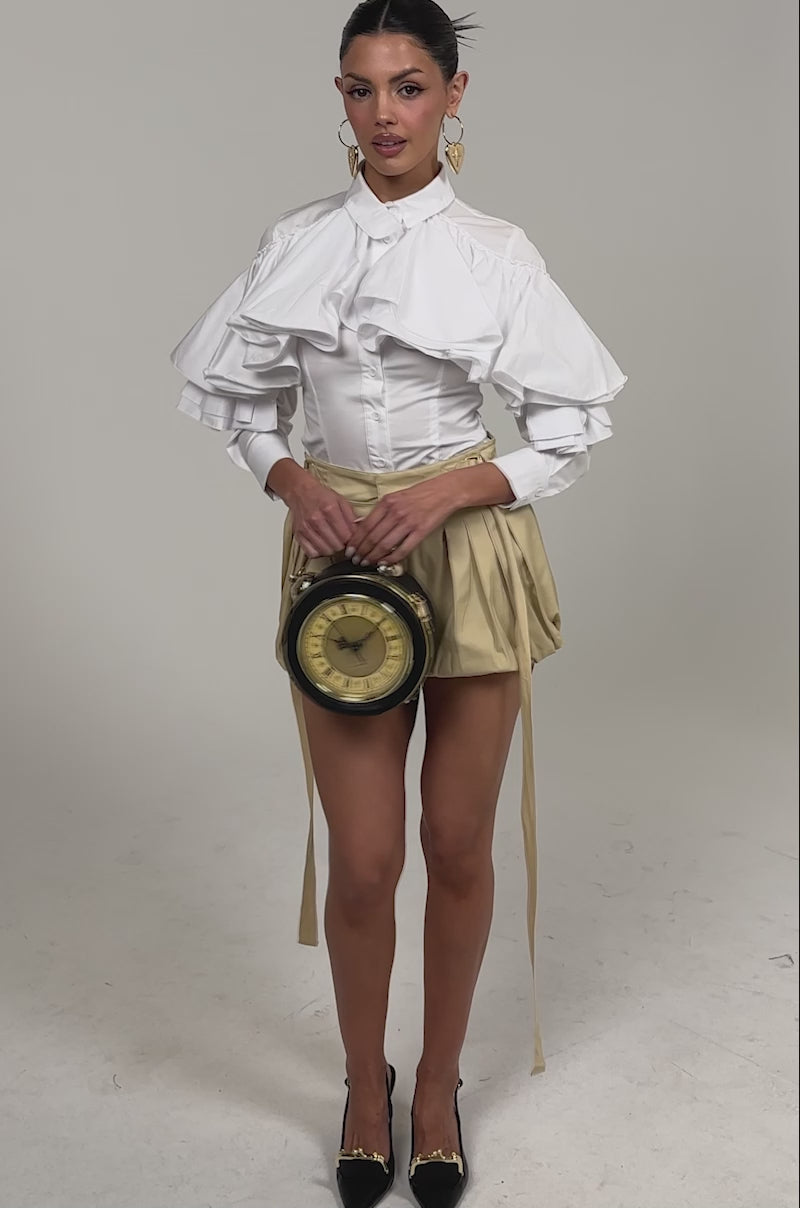 Video of woman modeling the SONIC PLEATED SHORT—beige, high-waisted bubble shorts with pleats and side ties—paired with a white ruffled blouse, black slingback heels, gold earrings, and a small black handbag against a plain light background.
