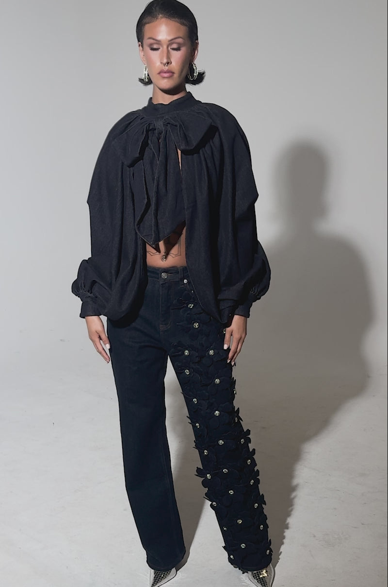 A video of a person posing in a dramatic dark outfit with a voluminous cape and FLORAL FIELDS APPLIQUE DENIM PANTS WITH RHINESTONES, standing against a plain gray background.