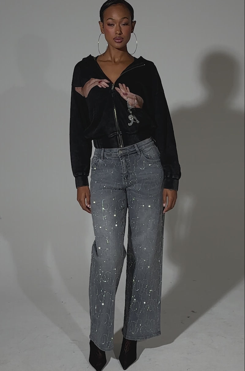 A video of a woman in the black GRAB MY HAND ZIP UP SWEATSHIRT with a hand graphic, paired with gray high-waisted patterned jeans, large hoop earrings, and black heeled boots against a plain light background.