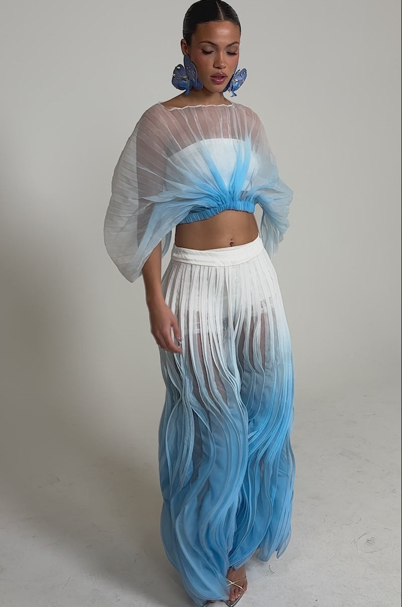 Video of woman modeling the IN THE SKY PALAZZO PANT—sheer ombre, wide-leg pants fading from white to blue with pleated, wavy textures—paired with large blue floral earrings.