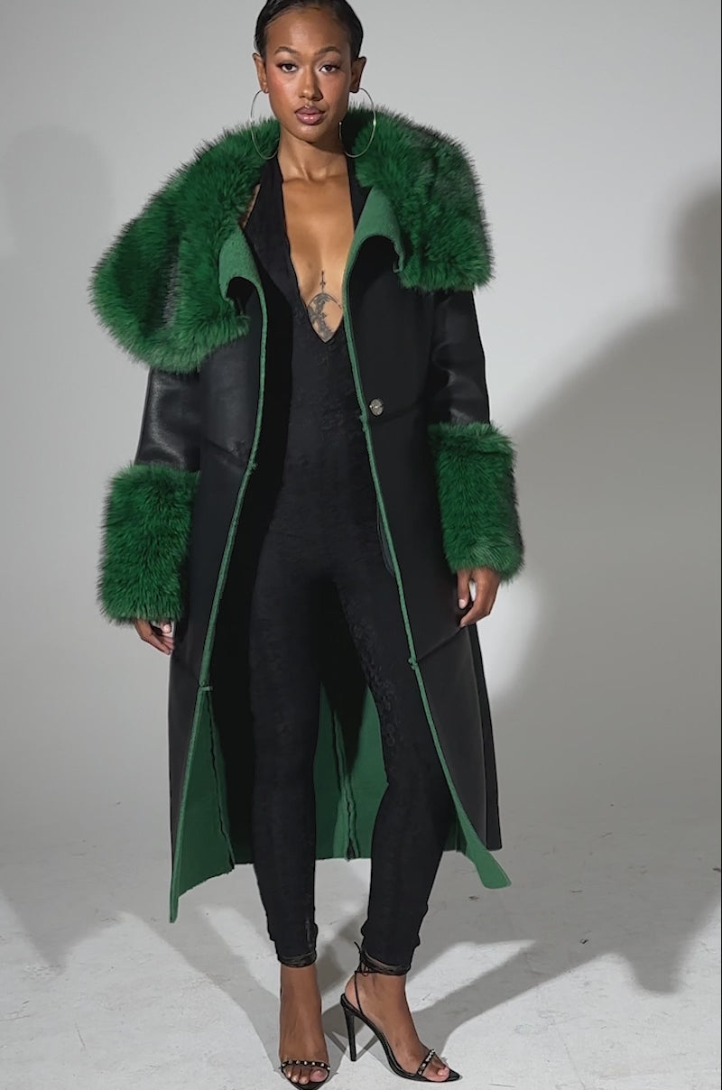 A video of a woman modeling the SEEING GREEN FUR TRIM TRENCH, featuring a bold emerald collar and cuffs, over a black lace jumpsuit with open-toed heels against a plain light background.