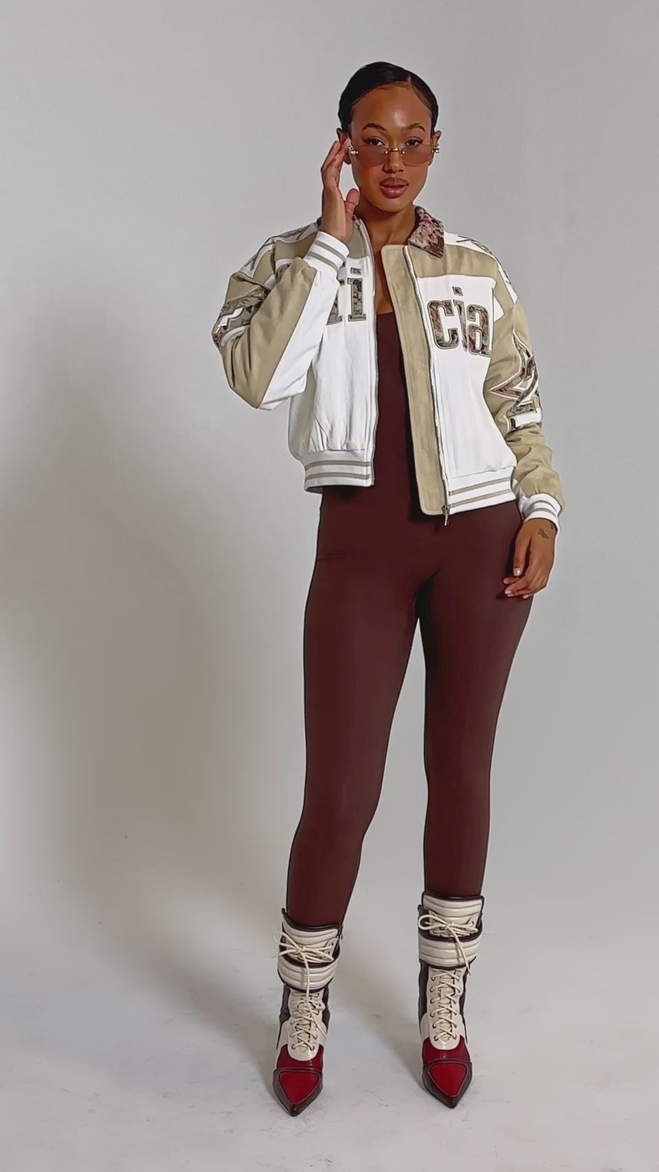 A video of a woman wearing the THAT GIRL SEAMLESS JUMPSUIT IN BROWN styled with sunglasses, a cream and beige varsity jacket, and lace-up heeled boots.