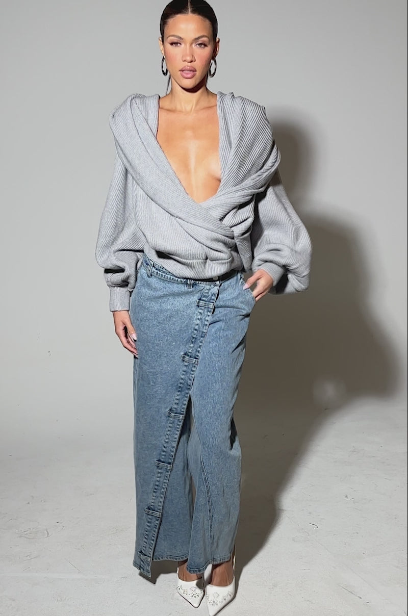 Video of woman modeling the COVERED UP RELAXED DENIM PANT—high-rise, wide-leg blue jeans with an asymmetrical front panel and buttons—styled with a gray sweater and white pointed-toe heeled mules for a modern, relaxed look.