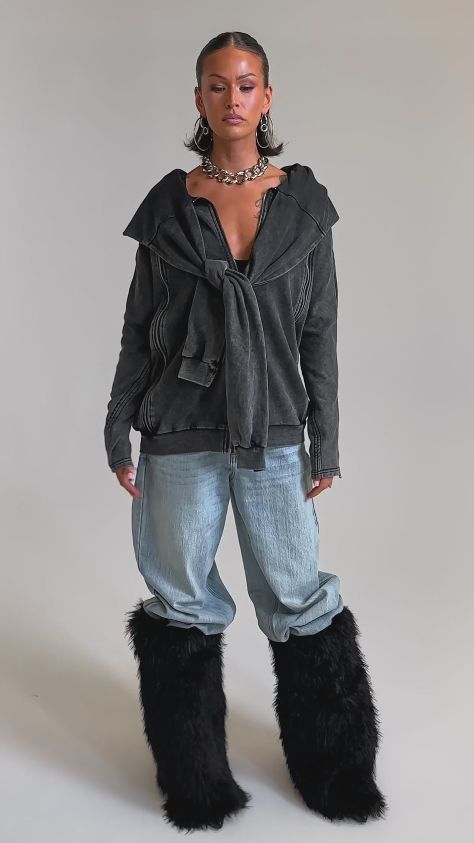 A video of a woman in the COOL GAL TIE SWEATSHIRT in dark gray, pairing it with light blue baggy jeans and black fluffy knee-high boots. 