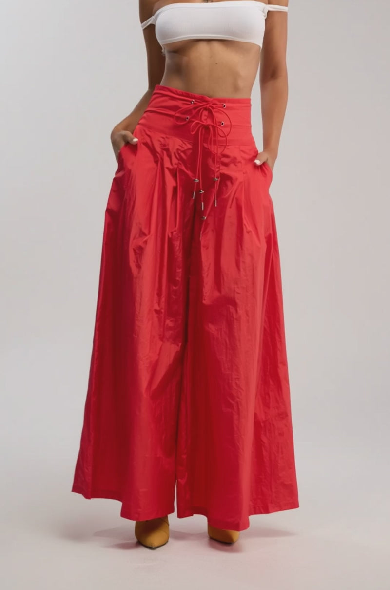 Video of woman modeling the DANCE THE NIGHT AWAY PALAZZO PANT IN RED, yellow sunglasses, yellow shoes, and a silver necklace standing with hands in pockets against a plain background.