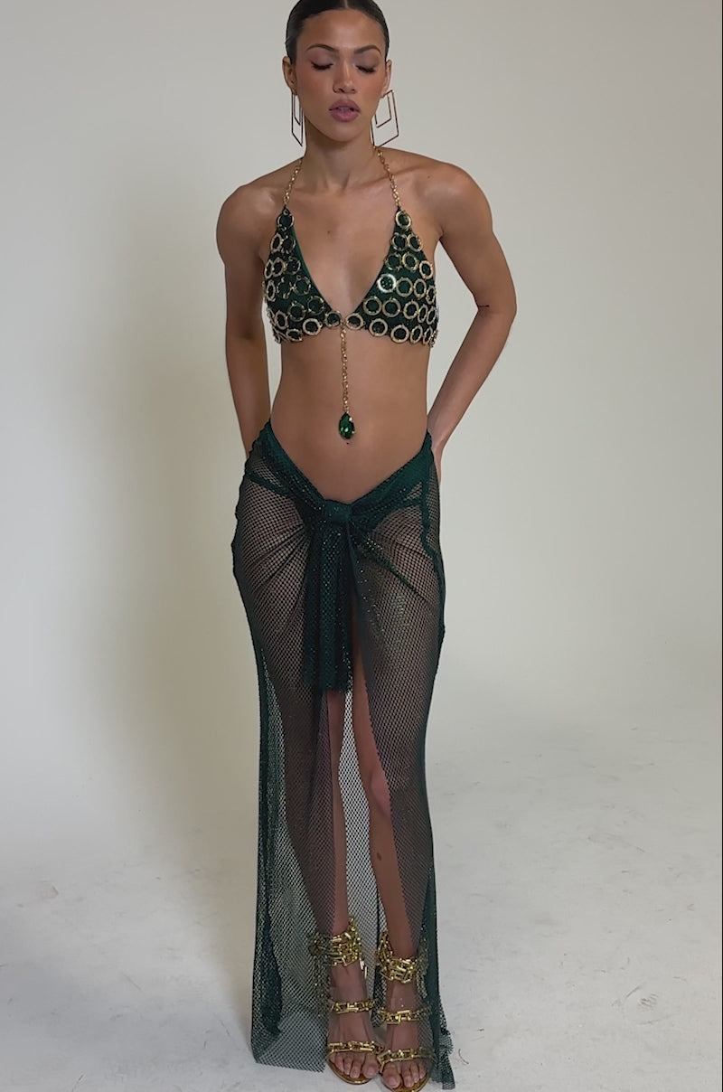 Video of woman modeling the MIAMI NIGHTS MESH MAXI SKIRT paired with a green patterned bikini top, gold jewelry, and gold high-heeled sandals for an elegant beachwear look against a plain light background.