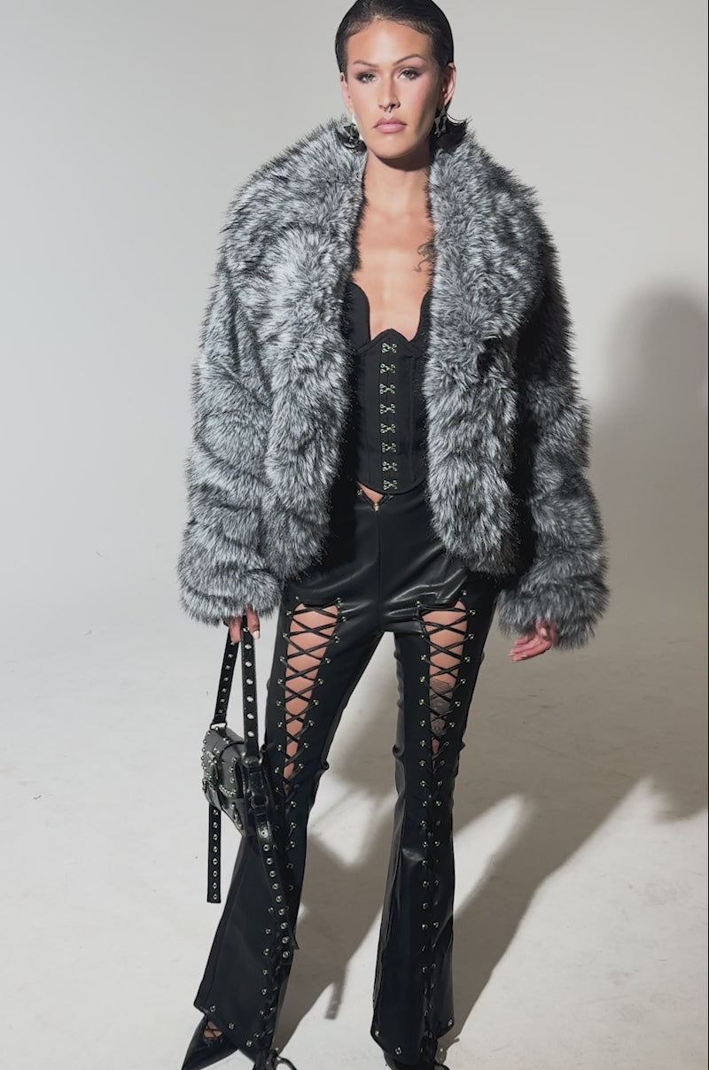 A video of a woman wearing the ELIXIR PANELED FAUX FUR IN GREY, black lace-up corset top, black lace-up leather pants, pointed black shoes, earrings, and holding a studded black handbag.