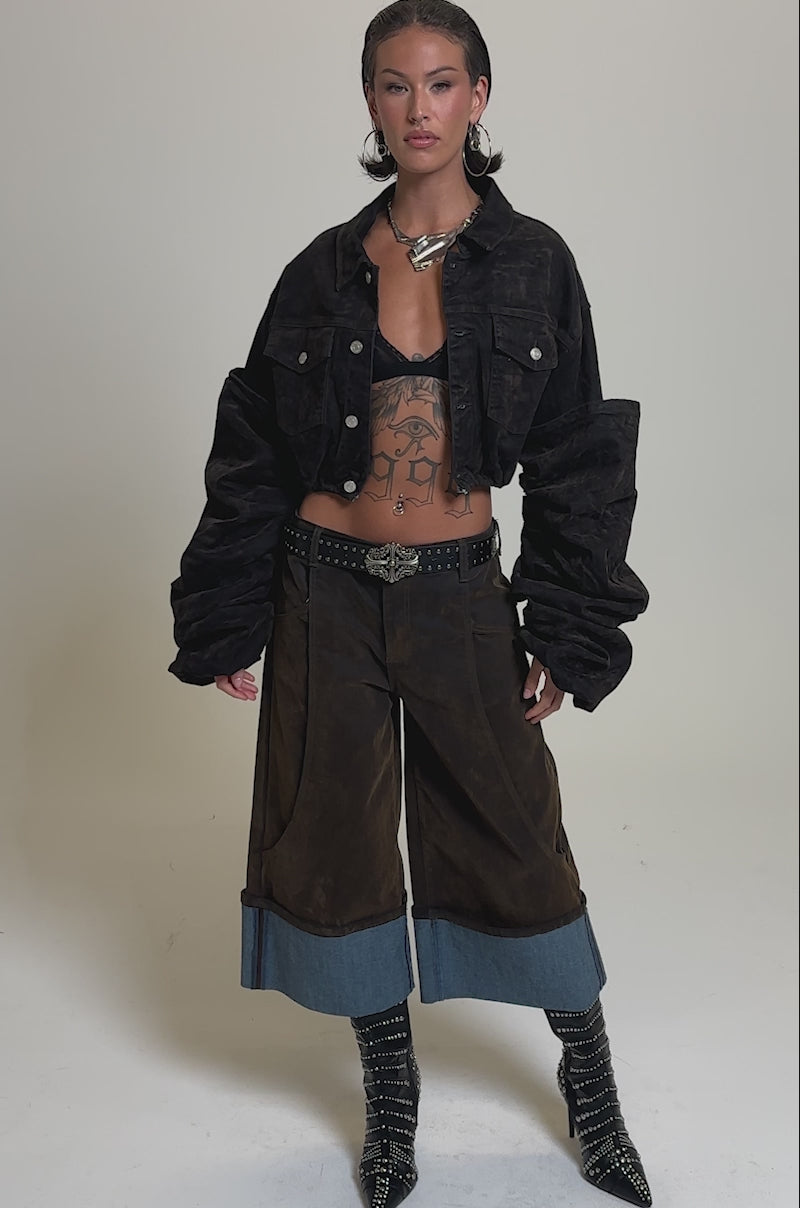 A video of a woman in the STACKED SLEEVE VELVET DENIM JACKET, paired with loose brown pants cuffed in denim, studded high-heeled boots, layered jewelry, and showing off her torso tattoos.