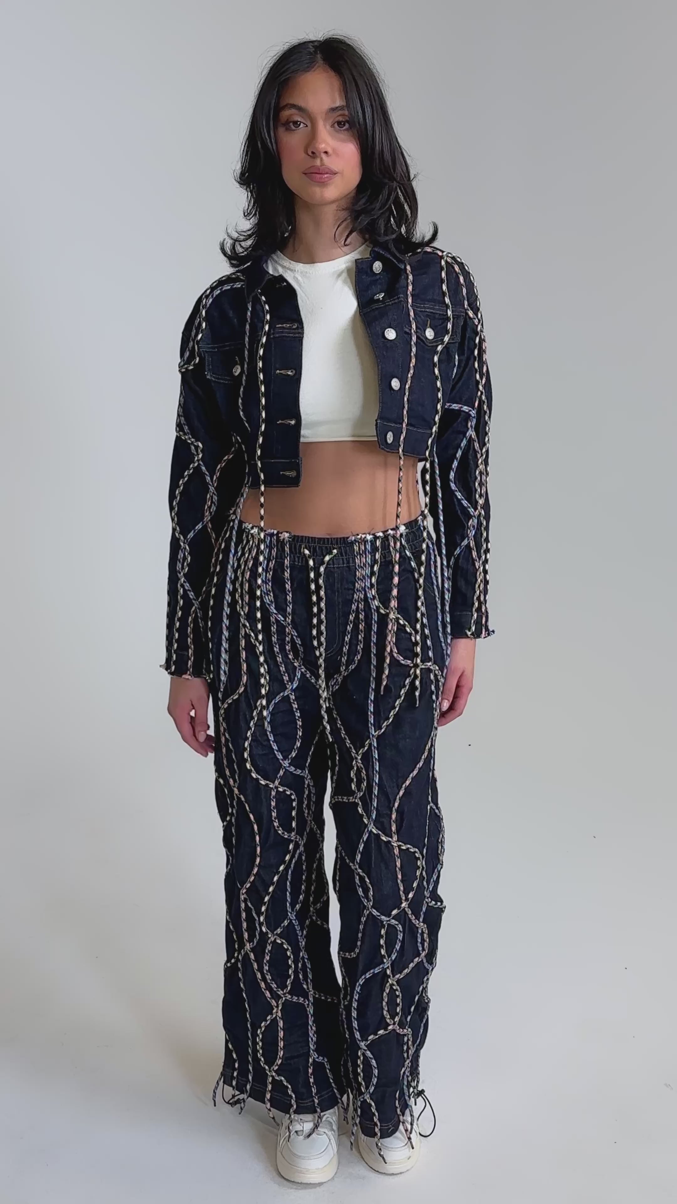 A video of a woman wearing the CRAZY FOR YOU DENIM JACKET with matching high-waisted chain-print pants, multicolor rope details, a white crop top, and white sneakers.