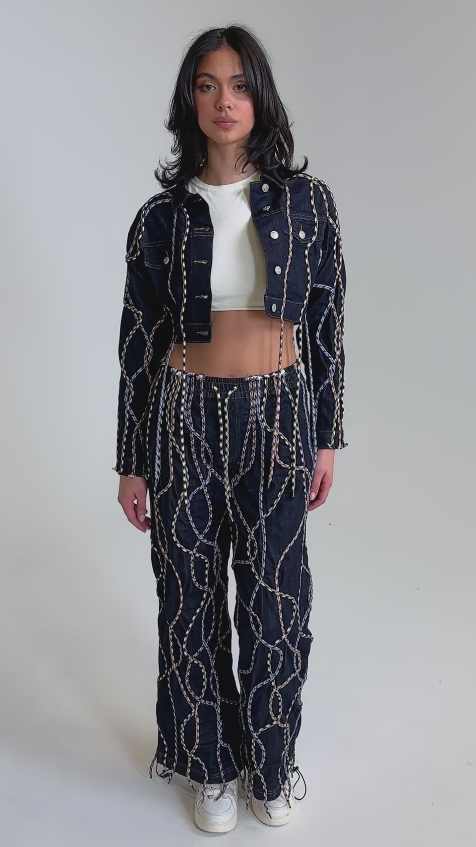 A video of a woman wearing the CRAZY FOR YOU DENIM JACKET with matching high-waisted chain-print pants, multicolor rope details, a white crop top, and white sneakers.