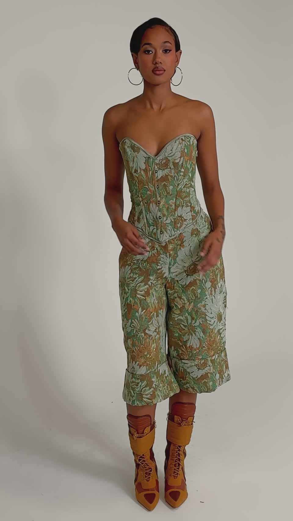 A video of a woman wearing the LOVERS FLORAL CORSET in green and orange with a strapless sweetheart neckline, paired with matching high-waisted jacquard culotte pants and gold strappy heels.