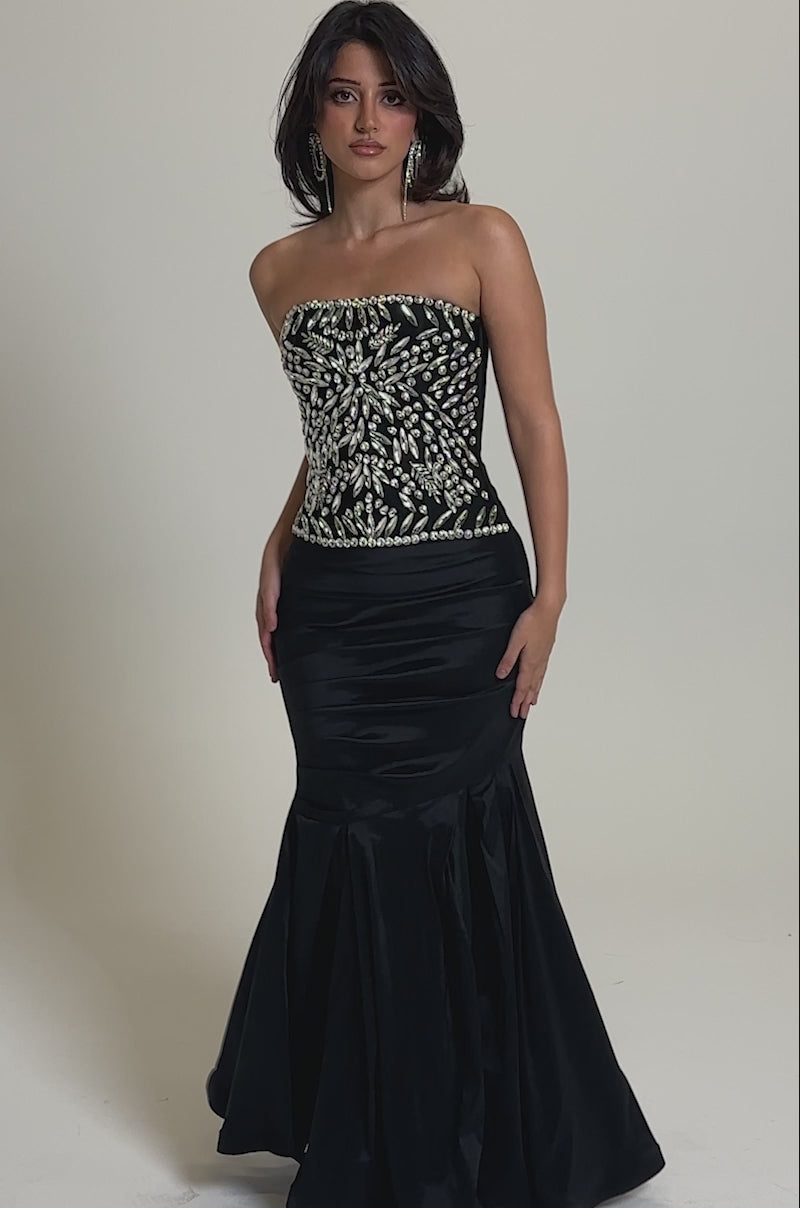 A video of a woman wearing the QUEEN OF THE NIGHT RHINESTONE CORSET black evening gown with a strapless neckline and rhinestone bodice.
