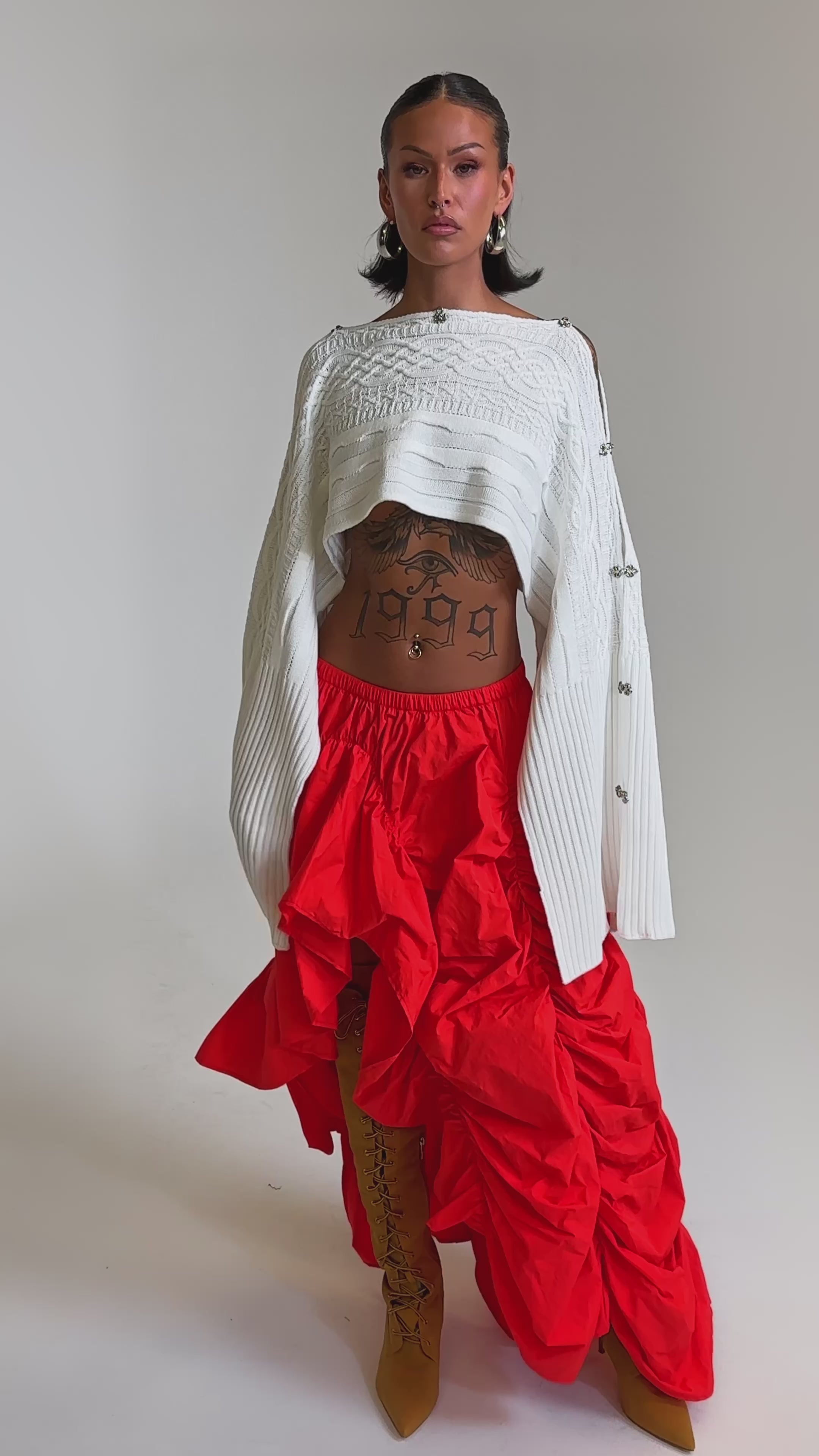 A video of a woman in a white, asymmetrical cropped sweater and the LETS CHANGE IT UP RUCHED MIDI SKIRT IN RED, paired with tan lace-up boots and hoop earrings. 