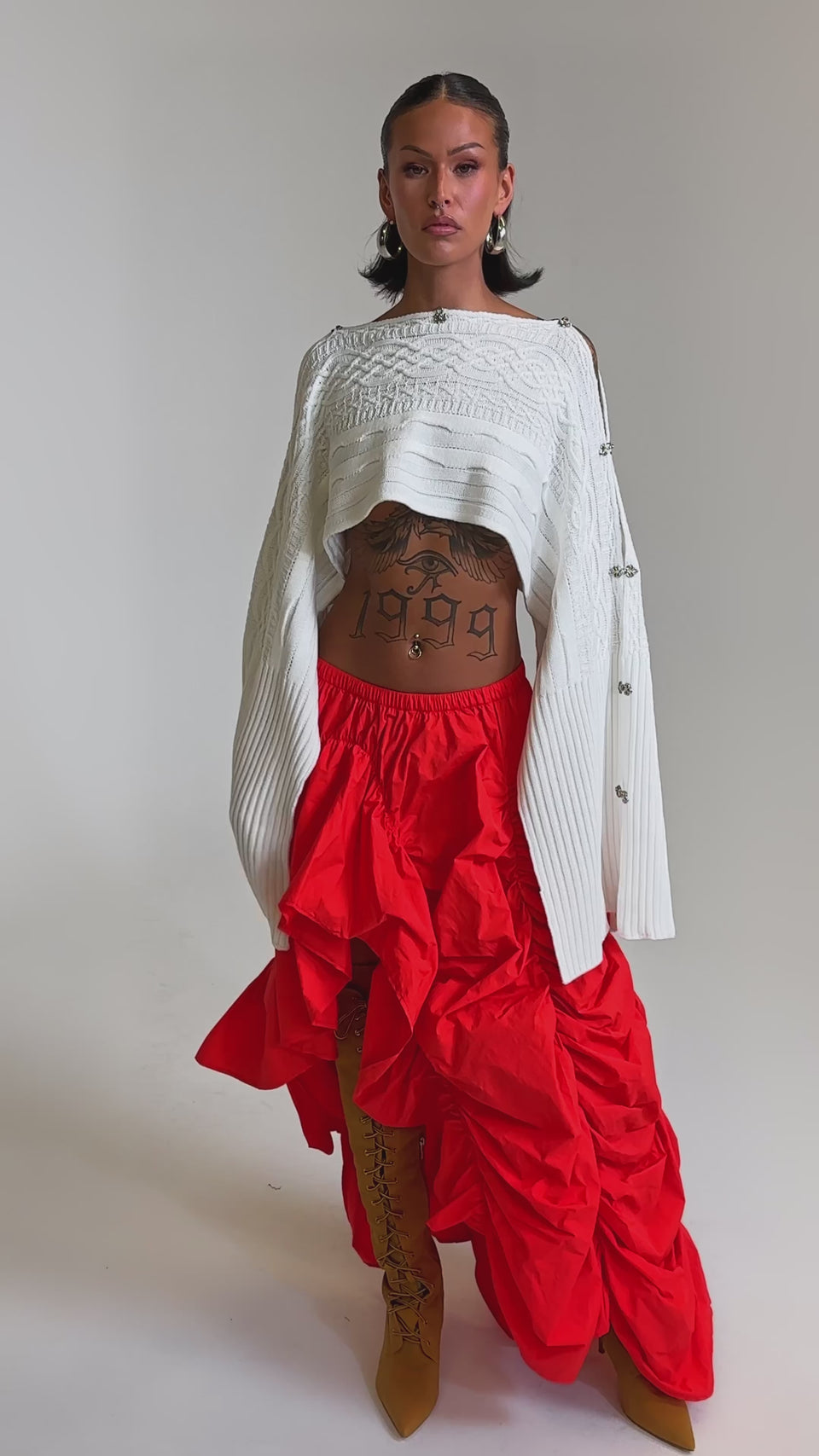 A video of a woman in a white, asymmetrical cropped sweater and the LETS CHANGE IT UP RUCHED MIDI SKIRT IN RED, paired with tan lace-up boots and hoop earrings. 