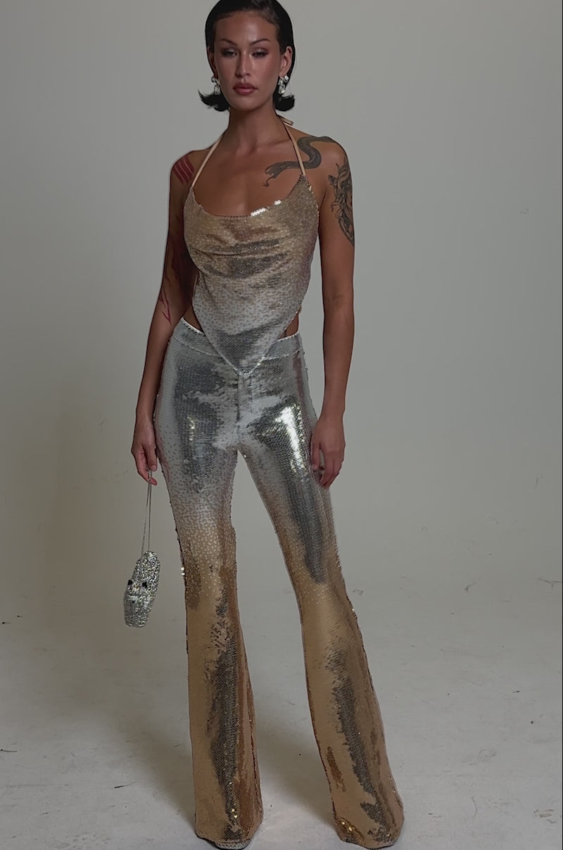 A video of a woman with tattoos and short, styled hair posing confidently in the GALAXY OMBRE SEQUIN HALTER TOP and matching flared pants, holding a sparkling handbag against a plain background.