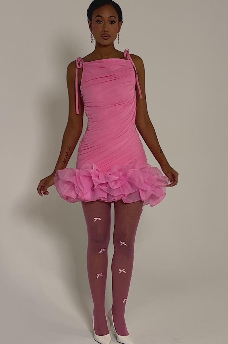 Video of woman modeling the PRINCESS POLLY PEPLUM MINI DRESS IN PINK, featuring a ruched bodice, organza ruffle hemline, tie straps, and pink fishnet stockings against a plain light background 
