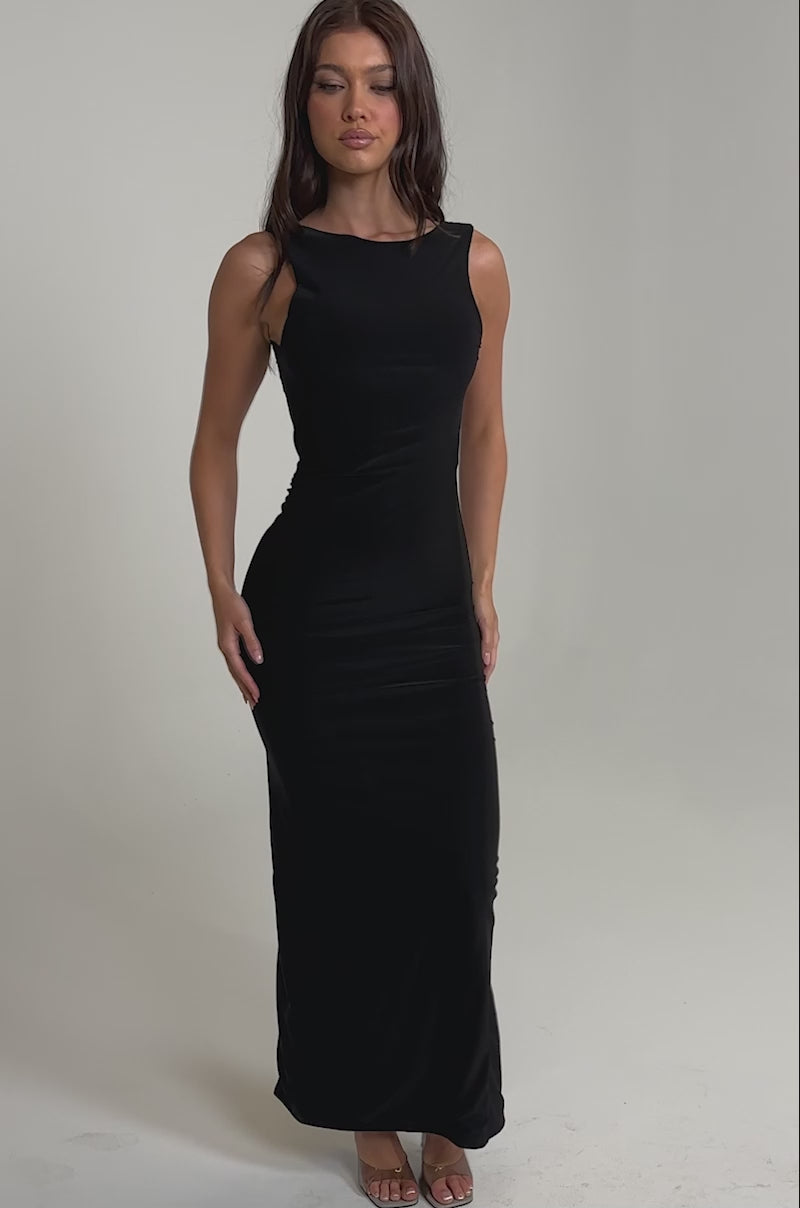Video of woman modeling the ON VACA MODE LOW BACK MAXI DRESS, a black maxi dress with a low-cut back and high slit, wearing clear heels 