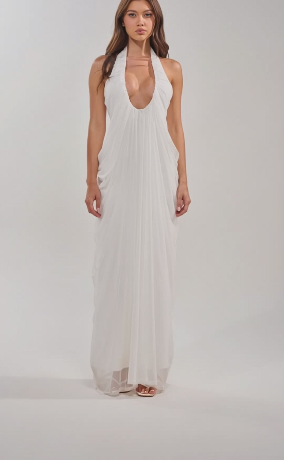 A video of a woman in the RAMONA PLEATED MAXI DRESS IN WHITE, a sleeveless, floor-length maxi dress featuring pleats, a plunging halter neckline, flowing draped fabric, and paired with white sandals.