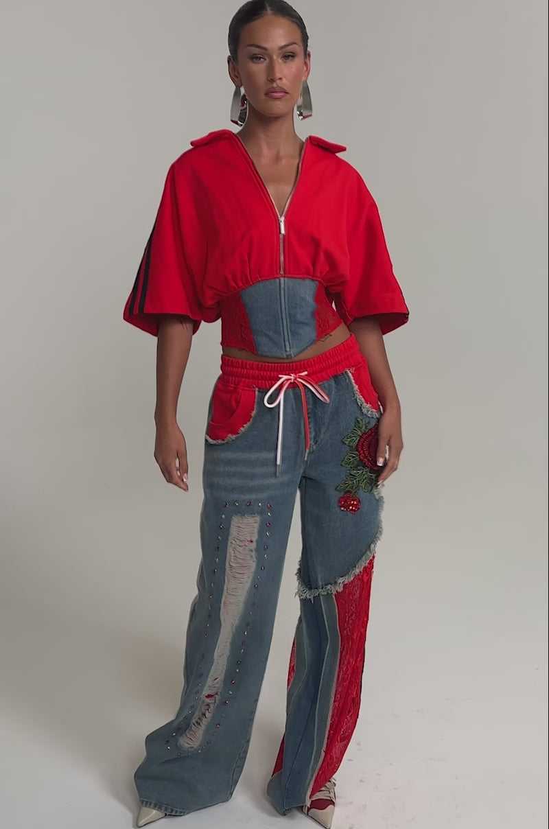 Video of woman modeling the GAME TIME JOGGER—high-waisted patchwork denim joggers with embroidery, red lace, and floral details—paired with a red cropped zipper jacket, statement earrings, and neutral shoes.