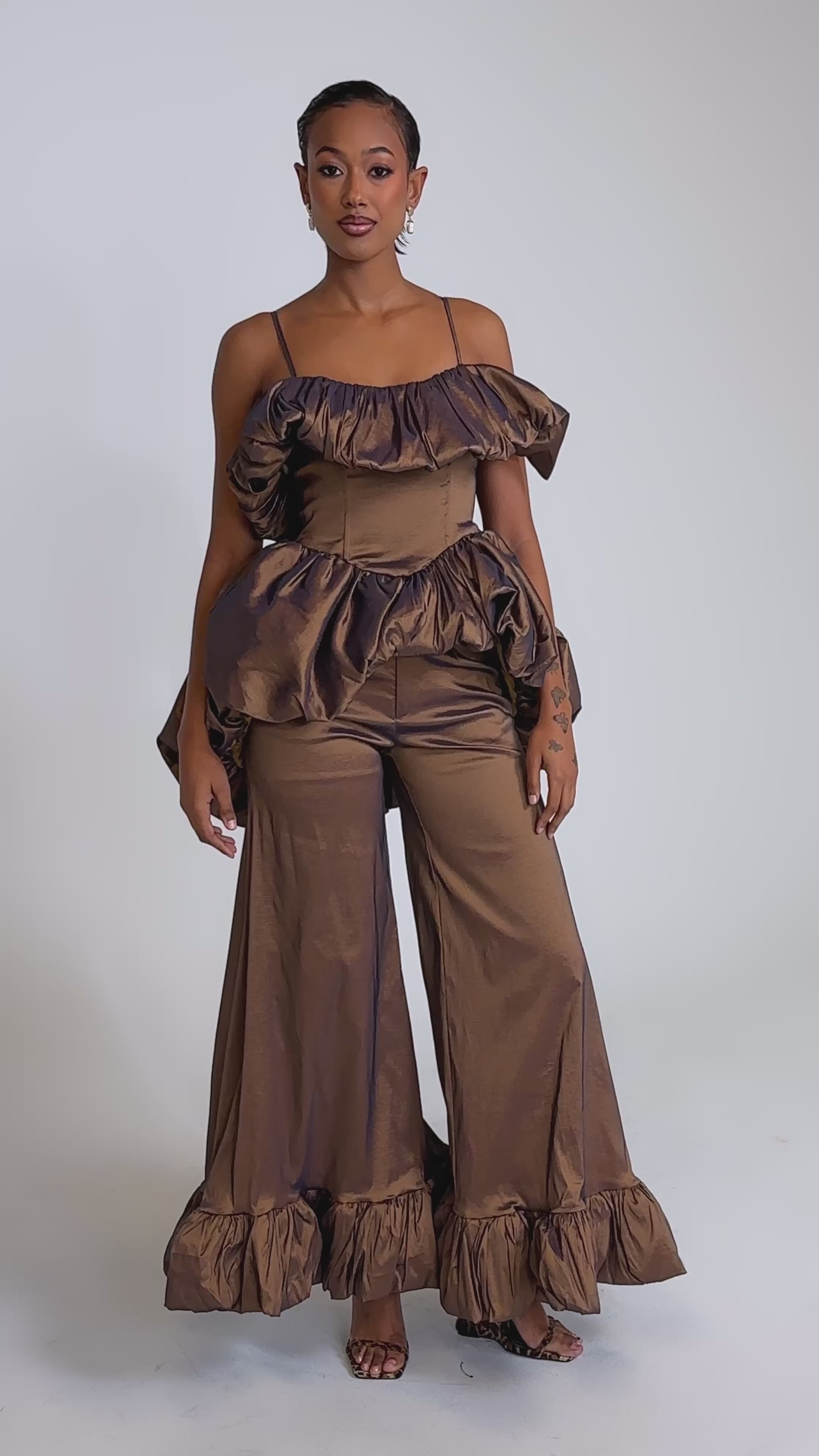 A video of a woman wearing the NOT TOO SIMPLE TROUSER—a shiny brown metallic taffeta wide-leg pant with ruffled hems—paired with a matching off-shoulder ruffled top, earrings, and heels against a plain background.