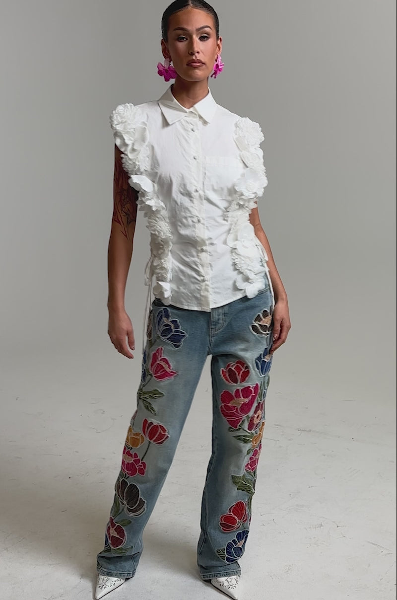 Video of woman modeling the CRAZY FOR YOU BUTTON DOWN, a cotton blouse with short sleeves and white 3D floral appliqué on the sides, paired with blue embroidered jeans. She accessorizes with pink flower earrings and displays a visible arm tattoo.