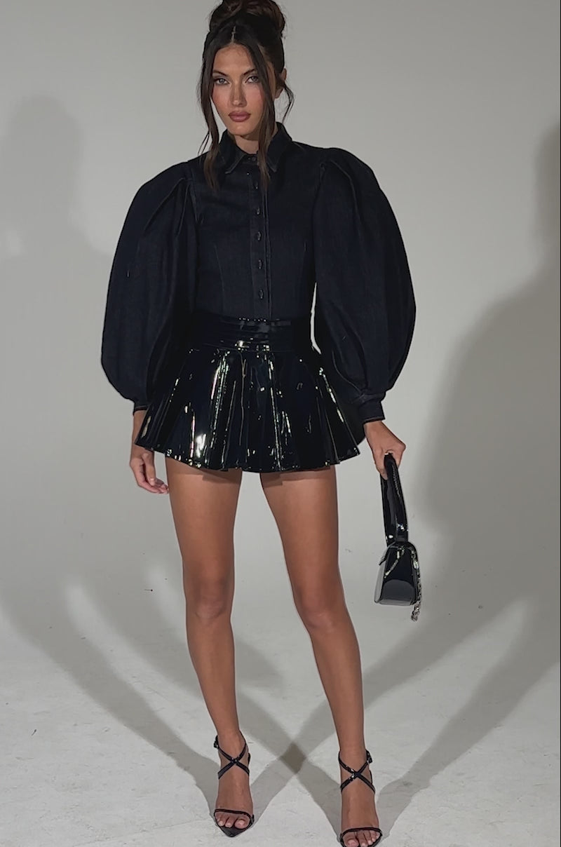 A video of a woman posing against a light background wearing a dark blouse with oversized puff sleeves, the SHE IS ON FIRE FAUX LEATHER MINI SKIRT IN NAVY, black strappy heels, and holding a small black handbag.