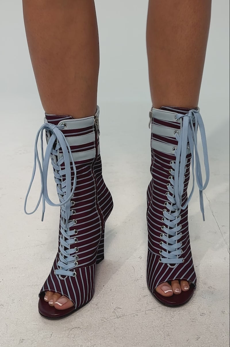 A video of a model wearing the AZALEA WANG WYATT BLUE MULTI STRIPE WEDGE BOOTIE which features a sporty wedge heel, open toe, maroon and white stripes, light blue laces, and a '42' patch at the top.