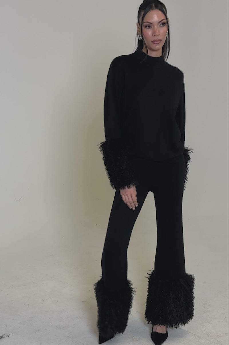 A video of a woman posing in the COZY VIBES SWEATER WITH FUR CUFF IN BLACK, styled with matching black pants and high heels, against a plain white background.