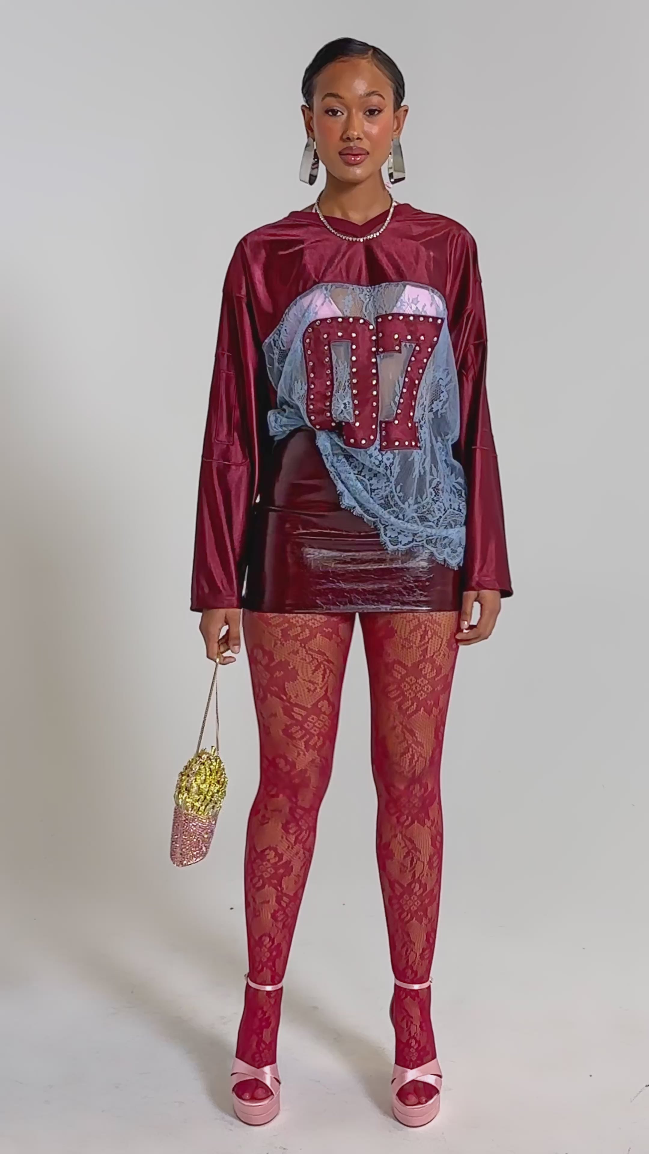 A video of a woman stands confidently in the sporty-chic WINNER JERSEY featuring “07” on the front, paired with red lace tights, pink platform heels, large earrings, and holding a rhinestone bucket with faux French fries.