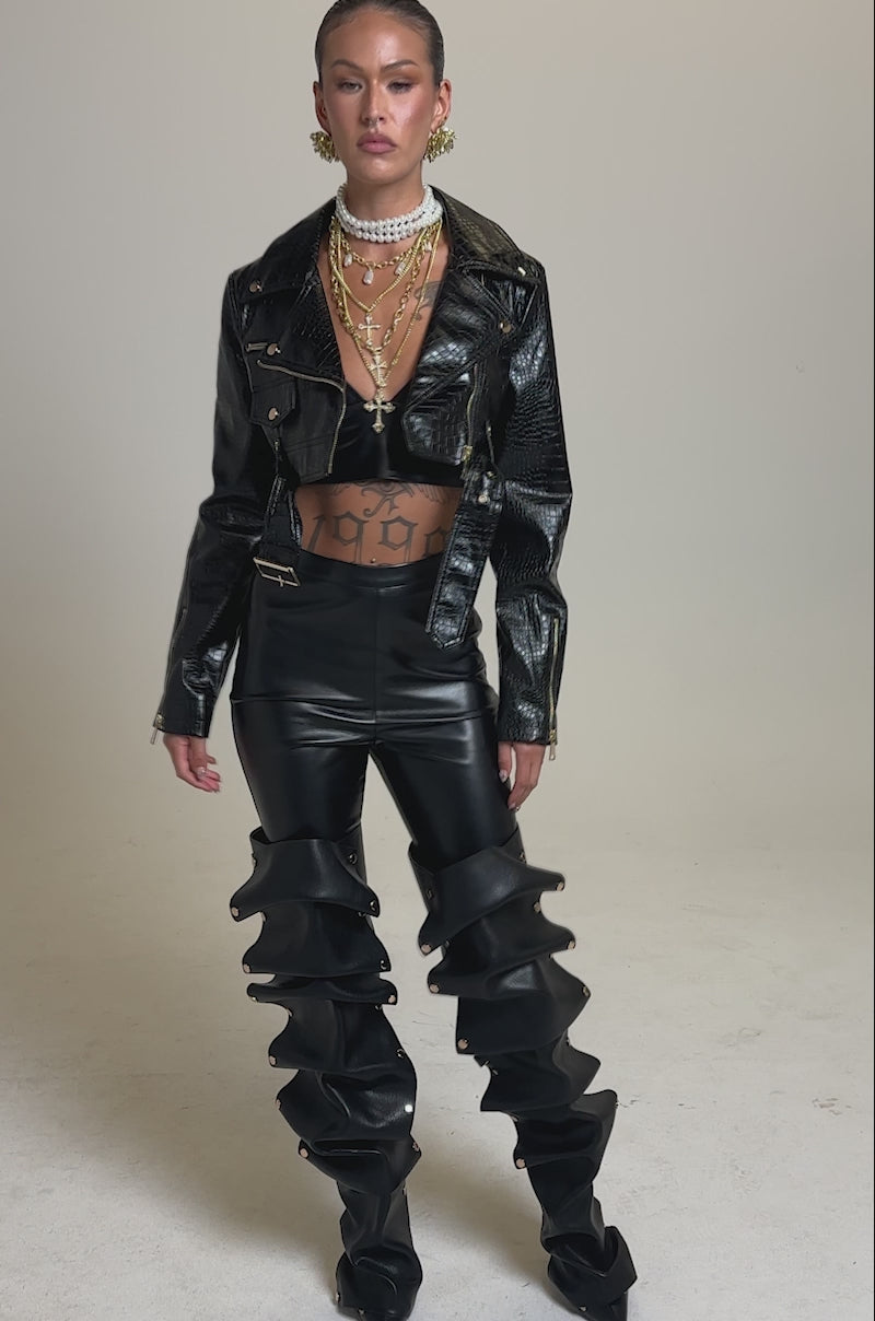 A video of a person posing confidently in the EVERYDAY MOTO IN BLACK CROC jacket, paired with high-waisted pants with dramatic ruffled legs, layered gold jewelry, and a sleek ponytail against a plain background.