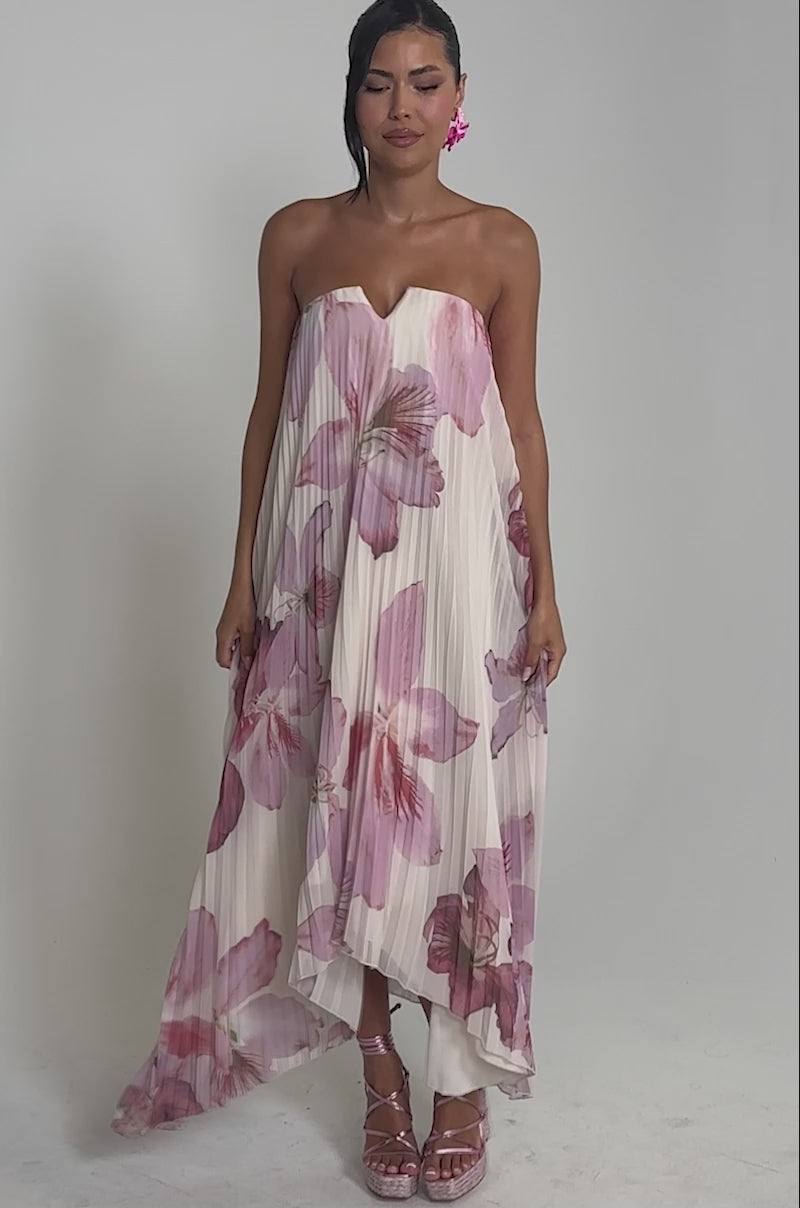 Video of woman modeling the JULIANA STRAPLESS FLORAL MAXI DRESS, featuring pink and purple pleats. She completes the look with large pink earrings, matching platform heels, and her dark hair styled to one side.