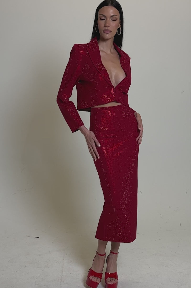 A video of woman wearing the ICED OUT RHINESTONE BLAZER—a sparkly, deep red, rhinestone blazer with long sleeves—paired with a matching high-waisted fitted midi skirt, long straight hair, and red platform heels.