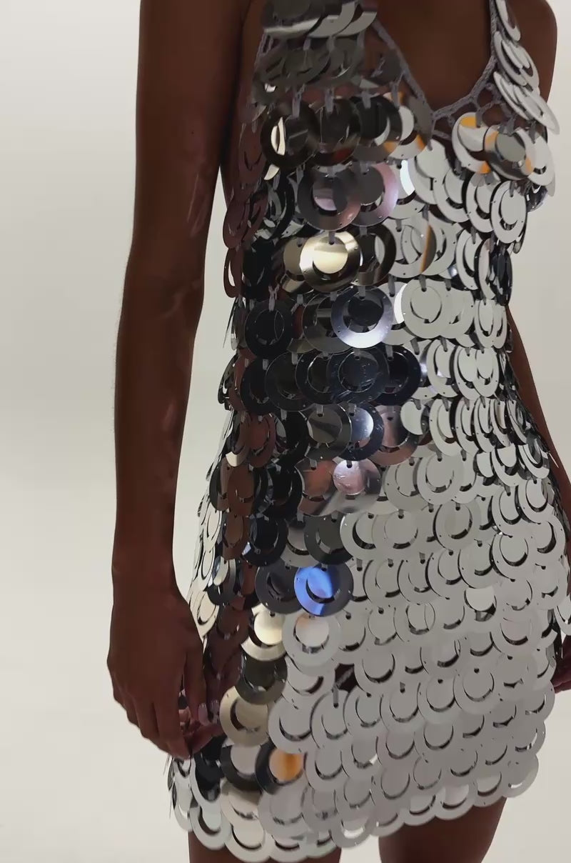 Video of woman modeling the SHIMMER AND SHAKE SEQUIN MINI DRESS IN SILVER, a sleeveless, deep V-neck halter mini dress with large shiny sequins. Paired with hoop earrings and slicked-back hair, it's perfect for bold festival looks against a simple backdrop.