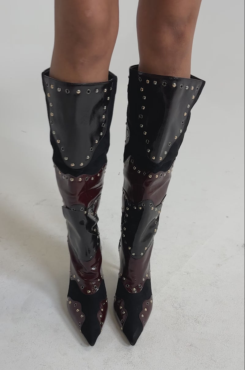 A video of a model wearing the AKIRA GOLD ENYAE MAHOGANY MULTI PATCHWORK STILETTO BOOTS—knee-high, pointed toe, black and burgundy faux leather with metallic studs.