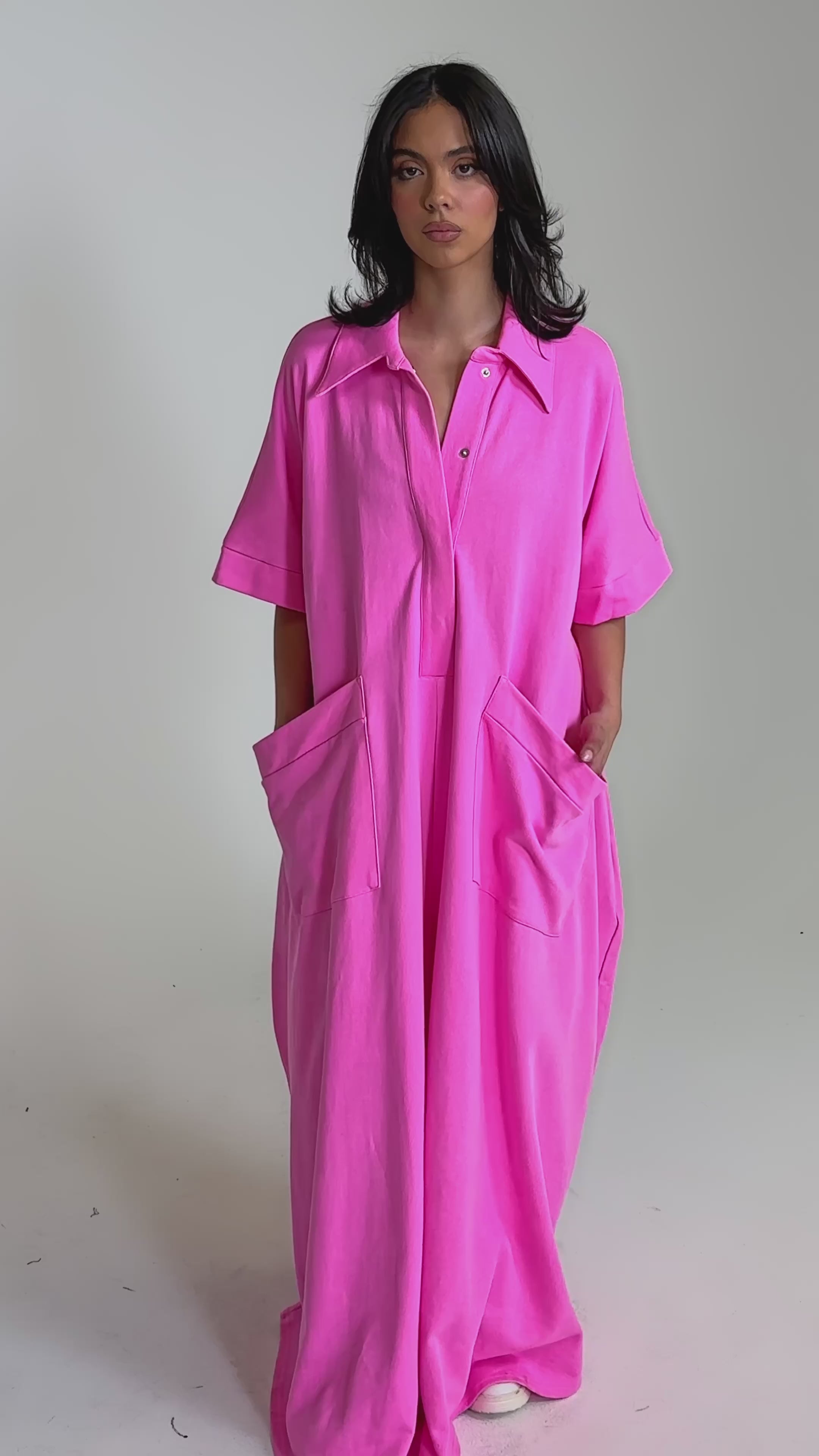 A video of a woman wearing the PRETTY IN PINK JUMPSUIT, an oversized bright pink one-piece with short sleeves, large front pockets, and a collar, standing against a plain light background.