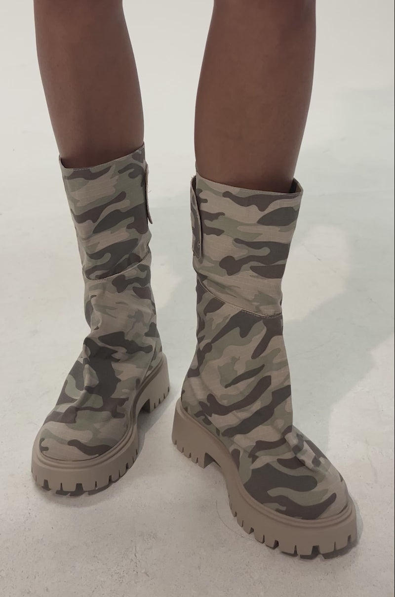 Video of woman modeling the AKIRA GOLD SPARK CAMO COMBAT BOOTIE, featuring a chunky heel and lug sole. Only their legs are visible