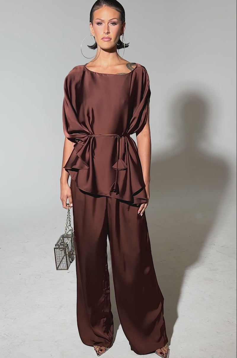 A video of a woman wearing the rust-colored HIGH SOCIETY BLOUSE with batwing sleeves and a waist tie, matching wide-leg pants, large hoop earrings, and carries a small metallic box handbag.