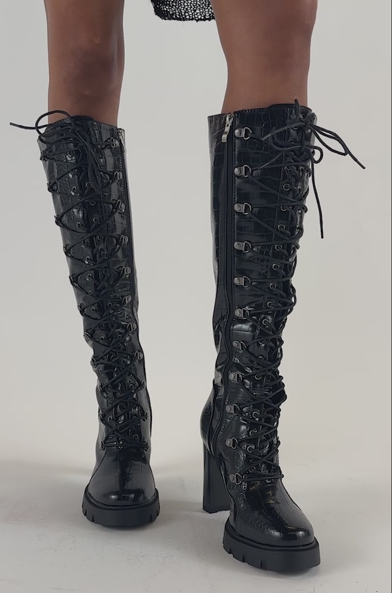Video of a woman modeling the AZALEA WANG AURIGA BLACK LACE UP BLOCK BOOT—tall, shiny black knee-high boots with high heels and a textured pattern.
