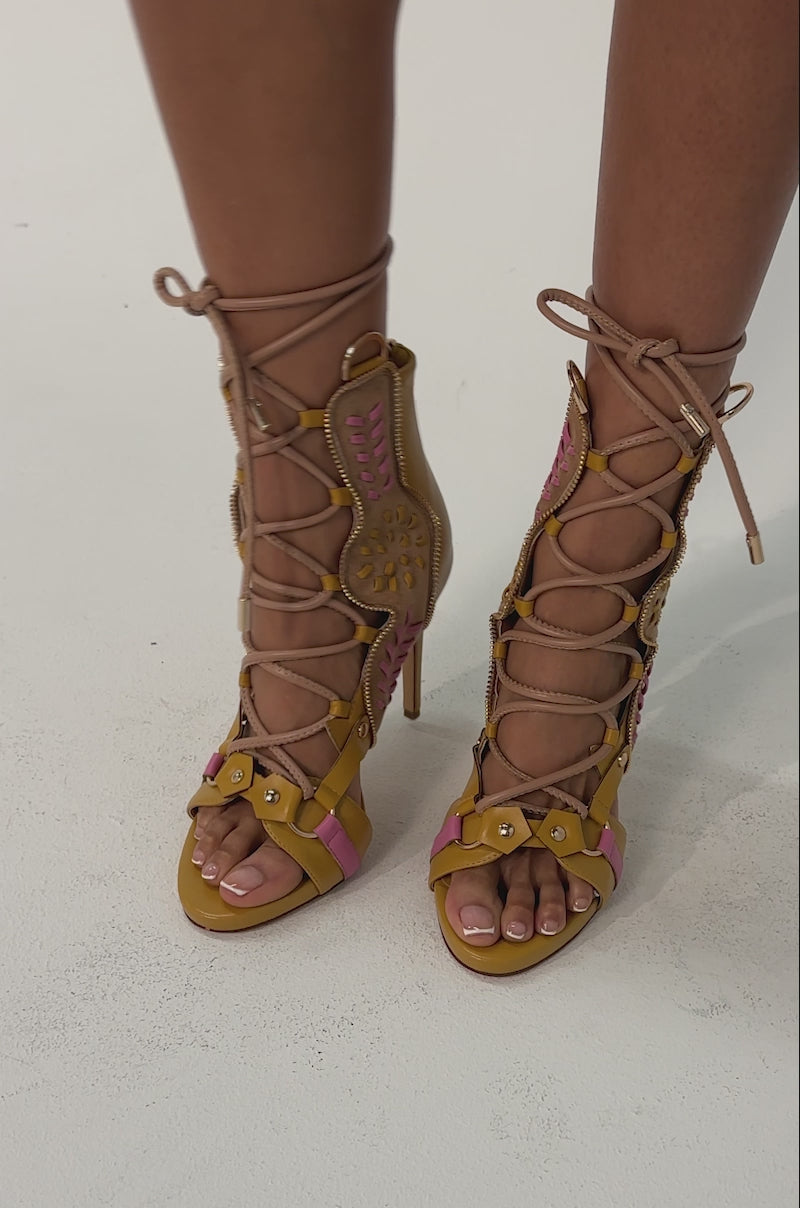 Video of woman modeling the AZALEA WANG SHAINIA MUSTARD MULTI STILETTO SANDAL, featuring tan hues with pink accents, cut-out details, and lace-up ankle ties. The stiletto heel offers a chic touch against a light background.