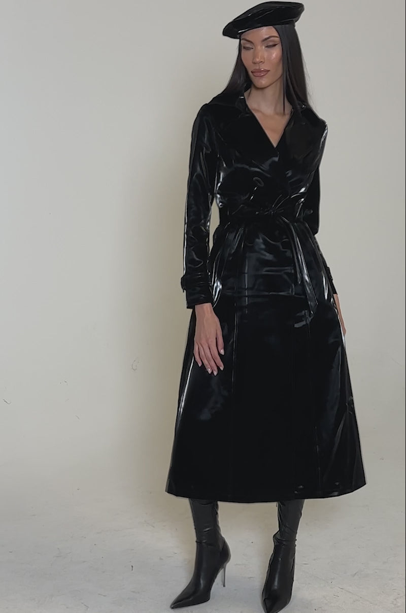 Video of woman modeling the SANDO LIQUID VELVET TRENCH IN BLACK, a glossy longline trench coat styled with a matching beret and knee-high heeled boots.
